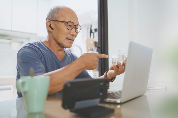 Chinese Senior male having video chat and holding medicine bottle for consult with doctor on laptop computer from home