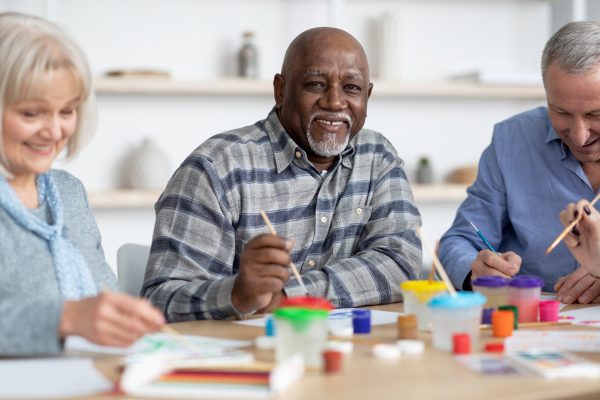 Leisure for senior people at retirement house concept. Happy senior african american man enjoying painting activity with his multiracial friends, holding brush and smiling at camera