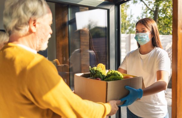 Young woman courier delivering purchases to elderly couple at doorway wearing mask and gloves